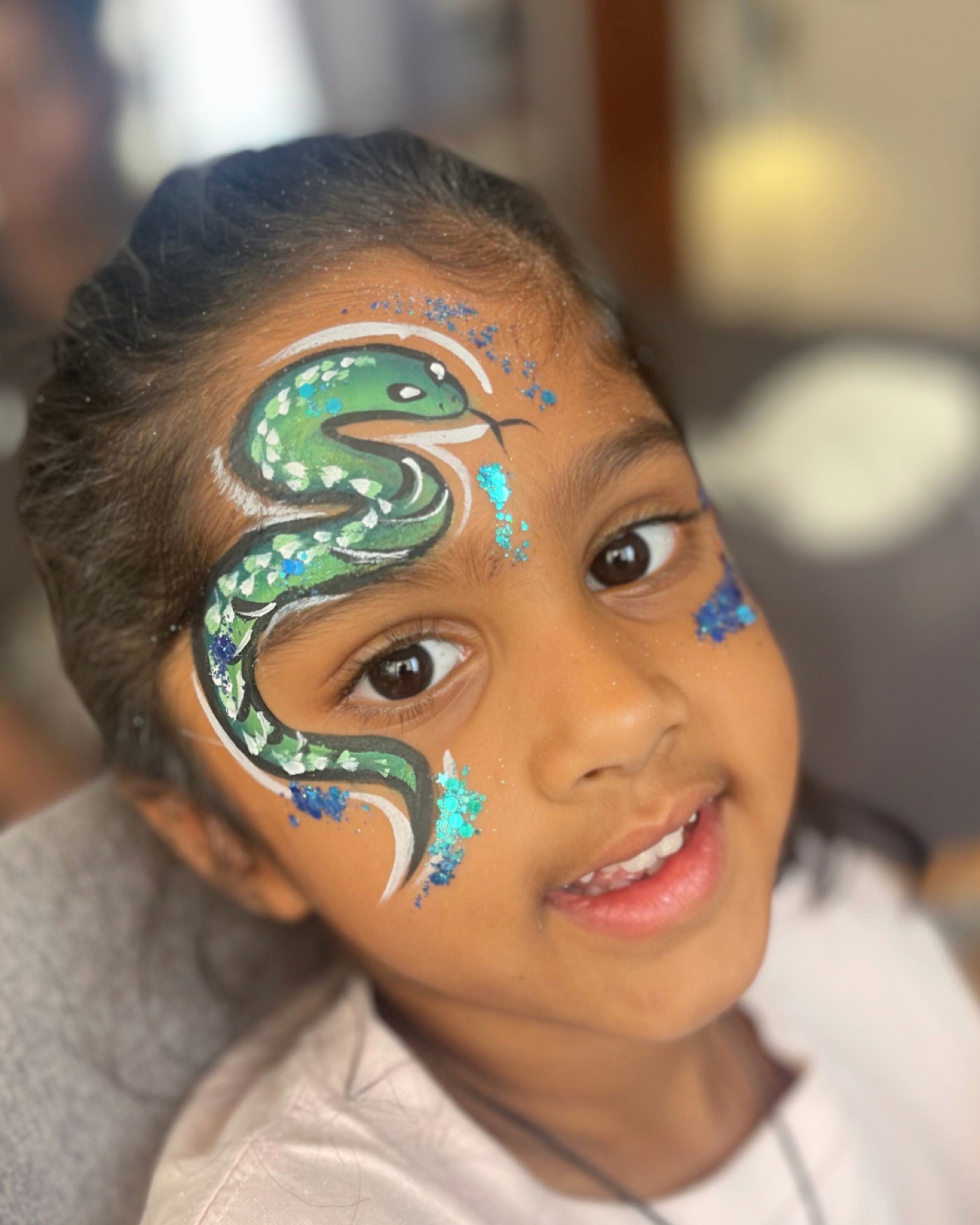 Smiling child showing an intricate green serpent face-paint design with blue glitter accents
